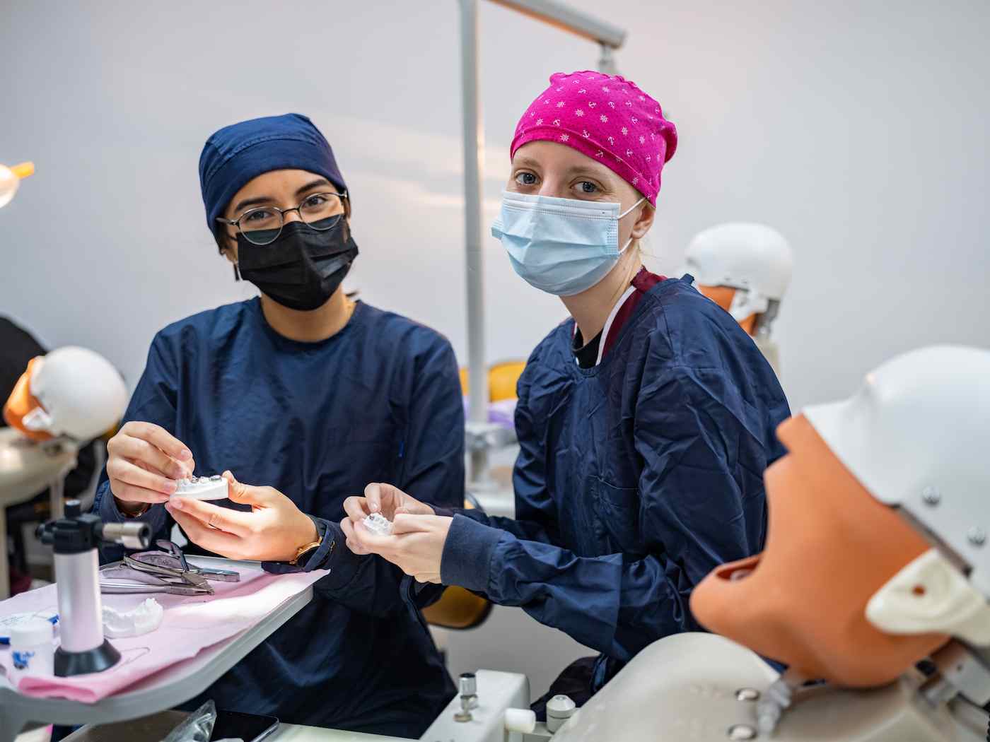 Dos estudiantes de odontología con mascarilla y uniforme quirúrgico practican con modelos dentales en un aula equipada con simuladores.